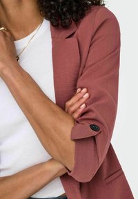Himbeerfarbener Leinenblazer mit strukturierter Oberfläche, hochgekrempelten Ärmeln und einem einzelnen schwarzen Knopfdetail am Bündchen, getragen über einem weißen Hemd.