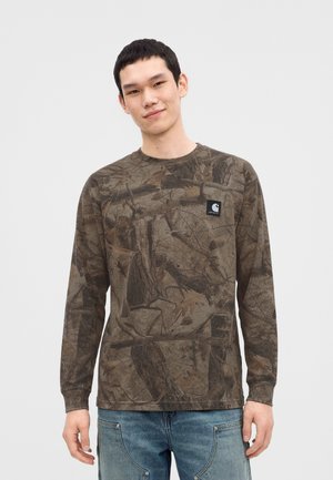 Giovane uomo che indossa una camicia mimetica marrone a maniche lunghe e jeans blu, in piedi davanti a uno sfondo chiaro e uniforme.