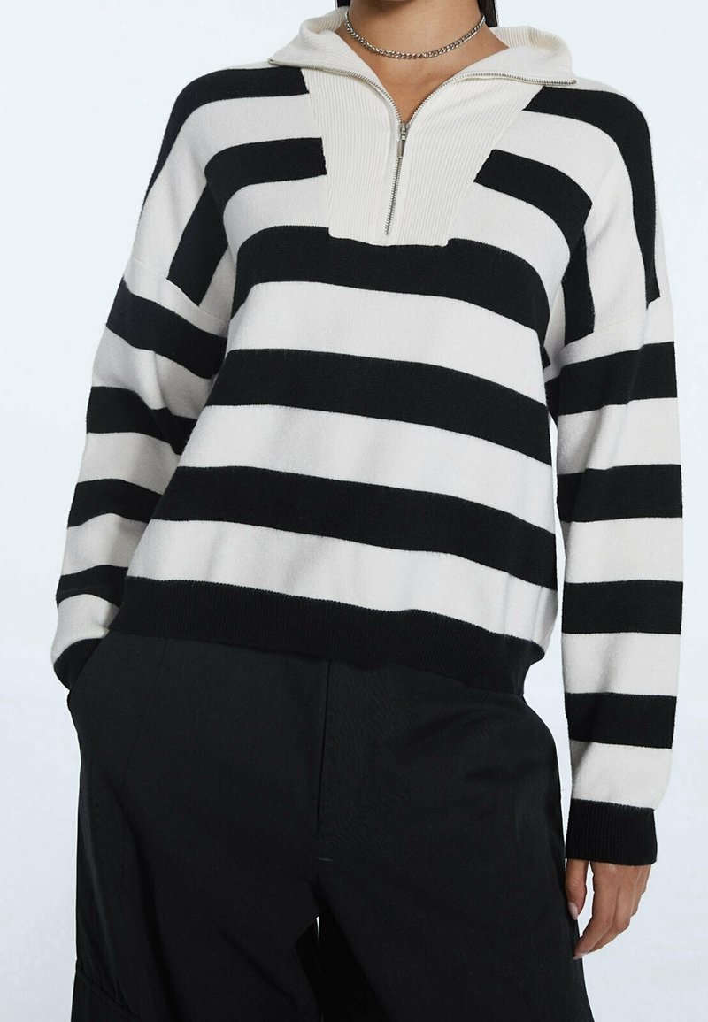 Pull en noir et blanc à rayures, fabriqué en tissu tricoté doux, avec une fermeture éclair quart de zip et un col côtelé. Design à coupe décontractée.