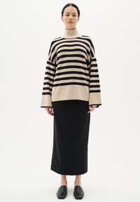 Pulover tricotat cu dungi în nuanțe de bej și negru, cu guler înalt, croială oversized, asortat cu o fustă maxi neagră și pantofi slip-on negri.