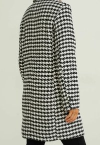 Manteau à motif pied-de-poule noir et blanc avec col, manches longues, fentes latérales et détails de boutons aux poignets.