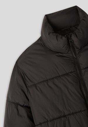 Primo piano di un giubbotto a sacco nero trapuntato con colletto alto e chiusura frontale con zip su uno sfondo chiaro.