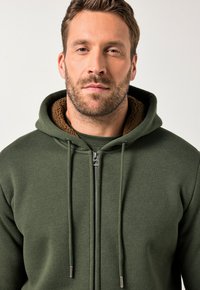 Olivgrüner Reißverschluss-Hoodie mit einer gemütlichen braunen Fleece-Innenausstattung, verstellbaren Kordeln und normaler Passform. Glatte Stofftextur, minimales Design.
