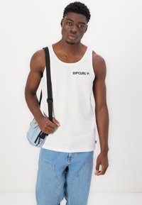 Top alb cu bretele, având logo-ul negru "RIP CURL", asortat cu o geantă albastră deschisă și blugi. Textură netedă, croială lejeră și design simplu.