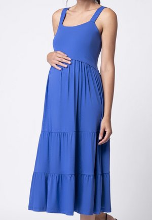 Femme enceinte portant une robe de maternité bleue sans manches, à étages, longueur genou, tenant son ventre d'une main.