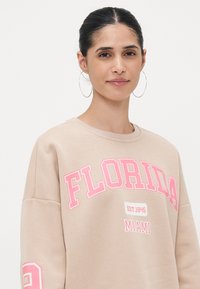 Felpa beige con la scritta "FLORIDA" e "MIAMI" stampata in rosa. Dettagli notevoli includono una vestibilità oversize e scollatura a girocollo con maniche rilassate.