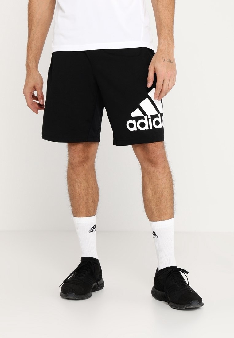 Pantalones cortos deportivos negros hechos de tejido suave, con un gran logo blanco de Adidas en el lado izquierdo, combinados con calcetines blancos hasta el tobillo y zapatillas negras.