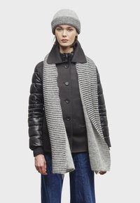 Donna che indossa un cappello di maglia grigio e una sciarpa rigata grigia sopra una giacca nera a maniche imbottite e pantaloni blu, in piedi su uno sfondo semplice.