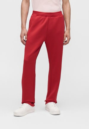 Hombre con pantalones deportivos rojos brillantes y zapatillas blancas, de pie frente a un fondo blanco simple.