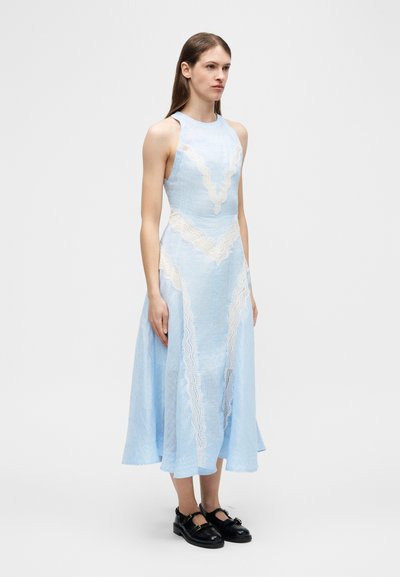 Γυναίκα που φοράει αμάνικο midi φόρεμα ανοιχτό μπλε με λευκές δαντέλες, όρθια, συνδυασμένο με μαύρα παπούτσια Mary Jane.