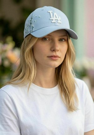 Blonde woman wearing a light blue cap with white floral embroidery and LA logo, and a plain white T-shirt, outdoors with blurred background.