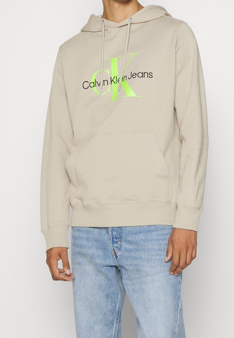Personne portant un sweat à capuche beige avec un logo "CK" vert néon et le texte noir "Calvin Klein Jeans", associée à un jean bleu clair, debout devant un fond blanc.