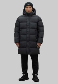 Abrigo de plumas negro con capucha, con un diseño acolchado, de longitud larga y con bolsillos laterales. Lleva puesto sobre pantalones y zapatos negros.