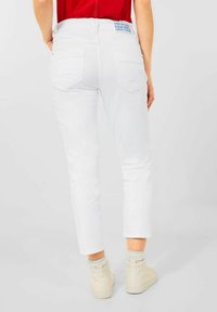 Jeans de mezclilla blanca con un ajuste slim, bolsillos traseros y una etiqueta azul en la cintura. Combinados con zapatillas altas de color crema claro.