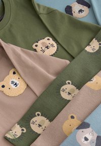 Ubrania dziecięce w stonowanych kolorach zielonym, beżowym i niebieskim, z teksturowanymi i nadrukowanymi twarzami niedźwiedzia i lwa jako wzorami dekoracyjnymi.