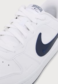 Sneaker bianca Nike con logo Swoosh blu navy, lacci bianchi e dettagli di cucitura visibili sulla parte superiore.