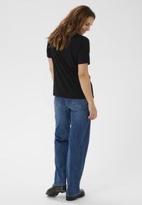 Hosszú barna hajú nő fekete rövid ujjú pólóban, kék egyenes szárú farmerral és fekete cipőben, állva, lefelé nézve, mosolyogva.