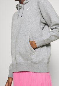 Grå huvtröja med framficka, dragsko och vitt Nike-märke. Buren över en plisserad rosa kjol. Mjukt tyg, ribbade muddar och fåll.