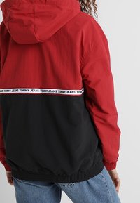 Osoba v červené a černé kapuci s logem "Tommy Jeans" na bílém pruhu na zádech, v kombinaci s modrými džíny.