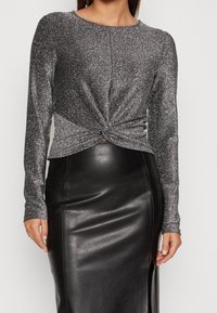 Glänsande silvrig långärmad topp med en vriden knutdetalj framtill, kombinerad med en svart fusk läder pennkjol med en sidoklyvning.