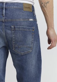 Blå denimjeans med en strukturert finish, med en rektangulær baklomme med en sikksakk-sømdetalj og en brun lærmerkelapp.