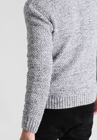 Mężczyzna ubrany w szary, teksturowany sweter dziany i czarne spodnie, pokazany od tyłu, z naciskiem na rękaw i bok swetra.