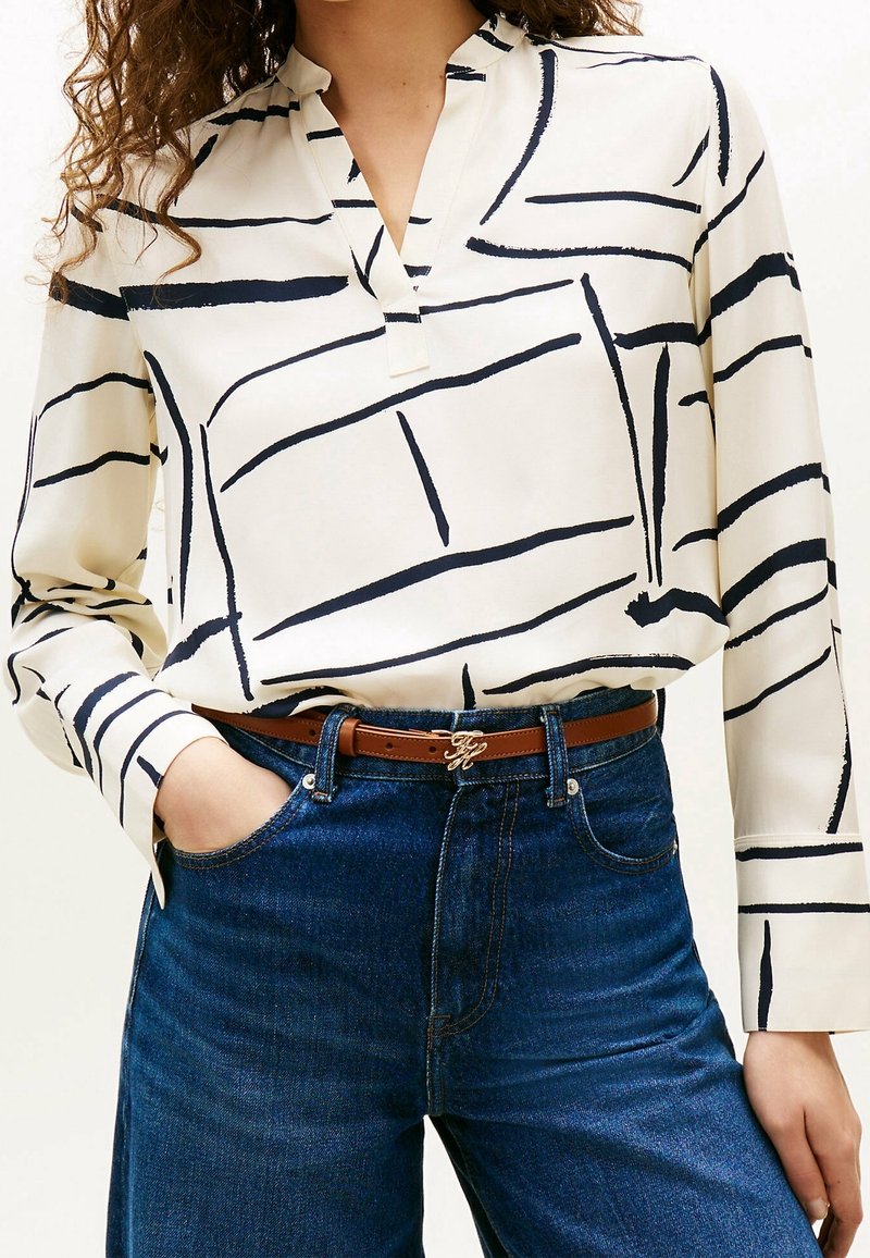 Femme portant un chemisier blanc avec des lignes abstraites noires, rentré dans un jean bleu taille haute avec une ceinture marron et une boucle dorée.