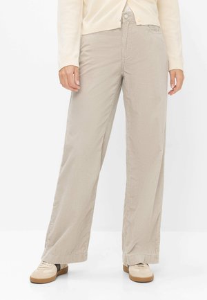 Frau trägt beige weite Cordhosen mit cremefarbenem Pullover und beigen Sneakers, steht vor weißem Hintergrund.