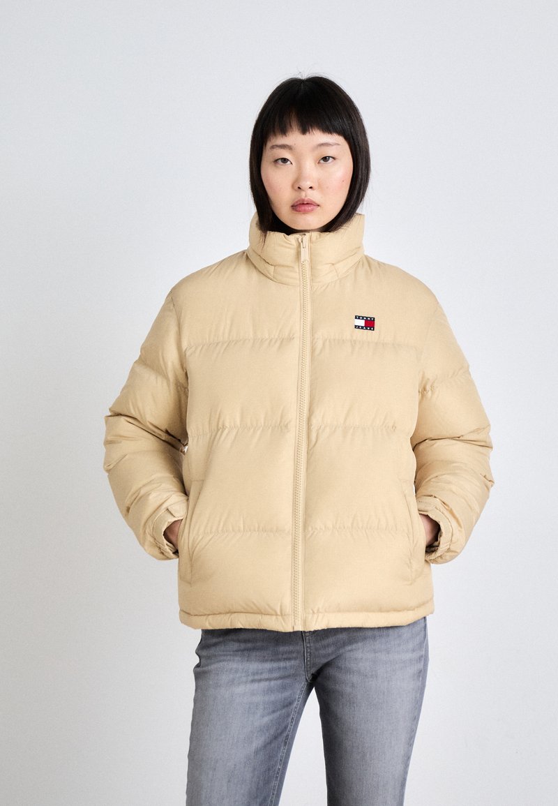 Beige Daunenjacke mit hohem Kragen, Reißverschluss vorne und genähten Paneelen. Verfügt über ein kleines Logo auf der linken Brust. Über einer Jeans getragen.