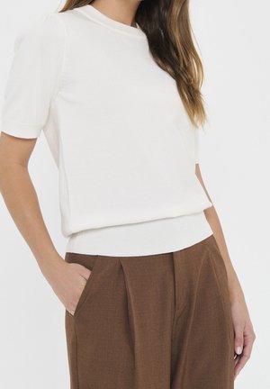 Femme portant un haut blanc à manches courtes rentré dans un pantalon plissé marron taille haute, les mains dans les poches.