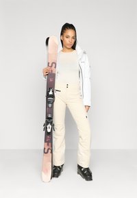 Σαφράνια σκι που διαθέτει λευκό μπουφάν, μπεζ παντελόνι σκι και μαύρες μπότες σκι. Κρατάει ένα καφέ και ροζ σκι με μαύρους συνδέσμους. Λείο υλικό.