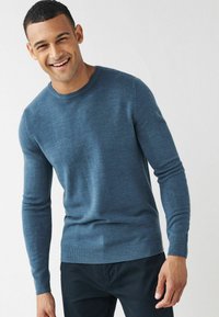 Jersey azul de hombre con mangas largas, escote redondeado, hecho de un suave tejido de punto, con dobladillo y puños de canalé. Lleva pantalones oscuros.