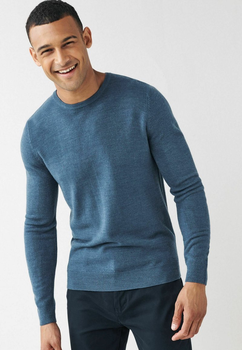Jersey azul de hombre con mangas largas, escote redondeado, hecho de un suave tejido de punto, con dobladillo y puños de canalé. Lleva pantalones oscuros.