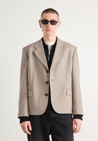 Blazer marrone chiaro con revers a punta, tre bottoni e tasche laterali, indossato sopra una camicia nera con zip e pantaloni scuri.