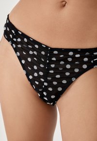Braguitas de bikini negras transparencias con lunares blancos. Presentan tejido fruncido y una cinturilla elástica suave. Vista en primer plano de la textura del material.