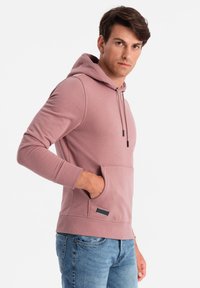 Mann trägt einen staubigen rosa Hoodie mit Fronttasche und blauen Jeans, steht mit einer Hand in der Tasche, leicht nach rechts gewandt.