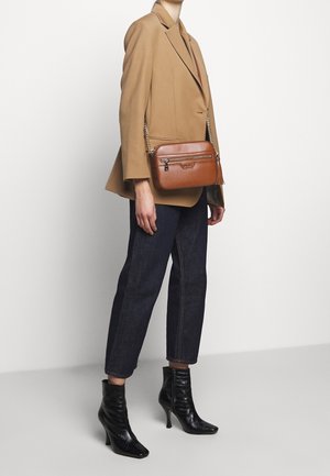 Borsa a tracolla in pelle marrone con design testurizzato, tasca con zip e tracolla a catena, indossata con un blazer beige e pantaloni in denim scuro.