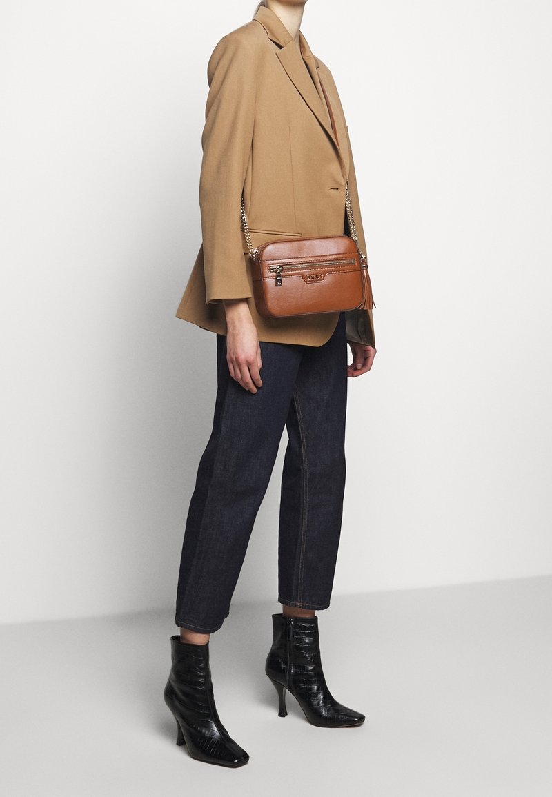 Brązowa skórzana torba na ramię z fakturowanym wzorem, kieszenią na zamek i łańcuszkiem, noszona z beżową marynarką i ciemnymi dżinsami.