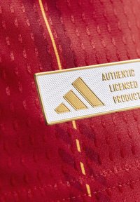 Crvena tkanina s teksturiranim finišem. Ima bijelu oznaku s zlatnim tekstom i tri dijagonalne pruge, koje označavaju autentičnost i licenciranje.