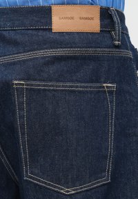 Gesäßtasche einer dunkelblauen Jeans aus Denim mit beigem Stich und einem braunen Lederpatch mit der Aufschrift "SAMSOE SAMSOE" am Bund.
