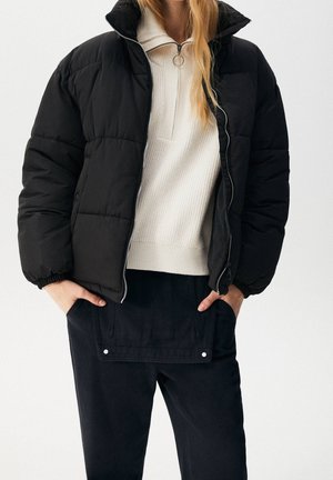 Person trägt eine schwarze Steppjacke über einem weißen Reißverschluss-Pullover und schwarze Hose, Hände in den Hosentaschen ruhend.