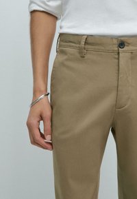 Khakihosen aus strukturiertem Stoff, mit einem vorderen Knopfverschluss, Gürtelschlaufen und einer Hand, die auf dem Bein ruht.