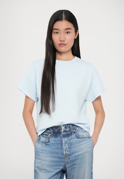 Lysblå bomulds-t-shirt med korte ærmer og rund hals. Båret med højtaljede, slidte denimjeans. Modellen står med hænderne i lommerne.