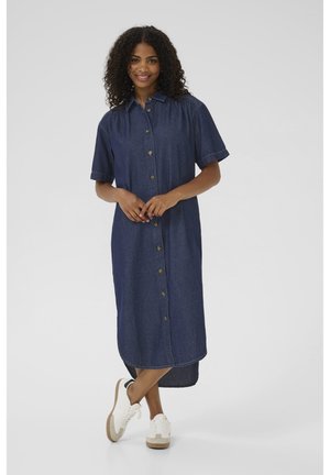 Robe en denim avec manches courtes, devant à boutons et longueur midi. Couleur bleu foncé avec des détails de couture subtils. Associée à des baskets blanches.