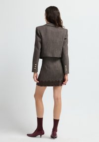 Bruine tweed cropped jas met gouden knoopaccenten, gecombineerd met een aansluitende bruine rok met een kantafwerking. Model draagt kastanjebruine sokken en schoenen.