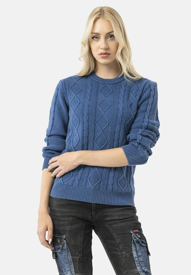 Pull en laine bleu à mailles torsadées avec un motif en losanges texturé, col rond, et poignets et ourlet côtelés ; associé à un pantalon sombre de style cargo.