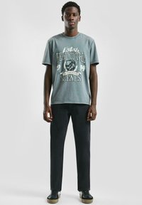 Młody mężczyzna stoi, ubrany w wyblakły zielony t-shirt z grafiką lwa i napisem, czarne spodnie i czarne tenisówki z beżowymi podeszwami na jednolitym tle.