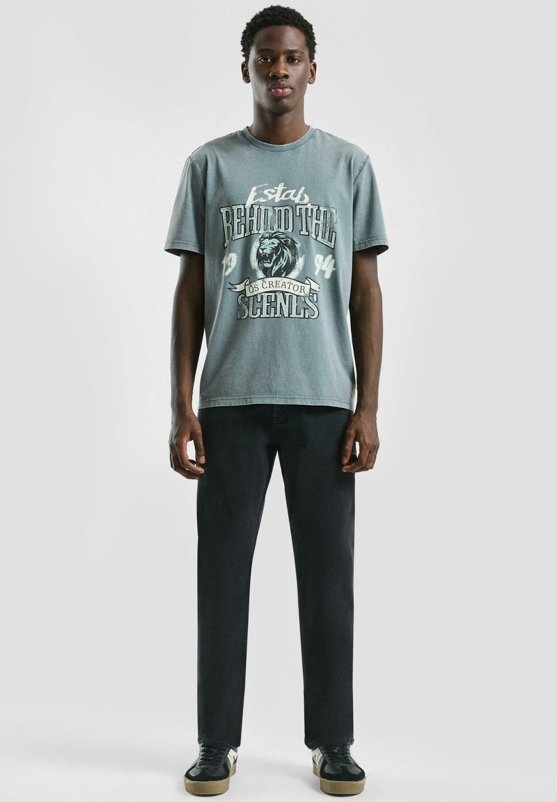 Młody mężczyzna stoi, ubrany w wyblakły zielony t-shirt z grafiką lwa i napisem, czarne spodnie i czarne tenisówki z beżowymi podeszwami na jednolitym tle.