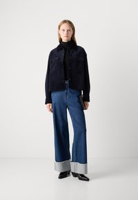 Veste cropped bleu marine avec fermeture à boutons et deux poches poitrine, associée à un jean oversized bleu avec des poignets en denim clair et des bottes noires.