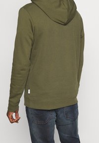 Grüne Baumwollkapuzenjacke mit einer Kordelzugkapuze, gerippten Bündchen und Saum. Kombiniert mit blauen Jeans mit genähten Gesäßtaschen.